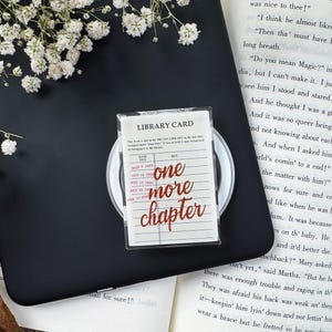 Puede incluir: Un soporte rectangular de acrílico transparente para teléfono con un diseño de ficha de biblioteca vintage y el texto "one more chapter" en letra roja. El accesorio está sobre un lector electrónico negro, encima de un libro abierto con flores.