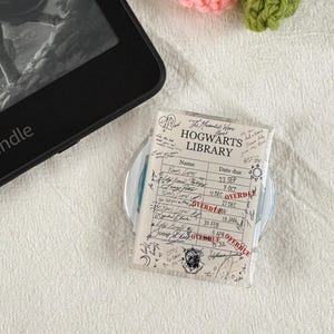 May include: A black e-reader displaying a book cover and a rectangular clear acrylic bookmark featuring a printed Hogwarts Library checkout card design with handwritten names, dates, and red overdue stamps, accented by a crocheted green vine with pink flowers.