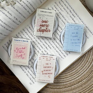 May include: Four rectangular acrylic keychains featuring printed designs resembling vintage library cards and guest checks. The designs include text such as one more chapter, buy me books & tell me I'm pretty, and the tortured readers department.