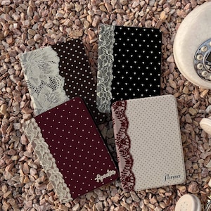 Puede incluir: Cuatro fundas rectangulares para pasaporte con patrones de lunares en negro, rojo oscuro y crema, cada una acentuada con una franja vertical de encaje intrincado. Dos fundas están personalizadas con los nombres Amelia y Florence en cursiva.