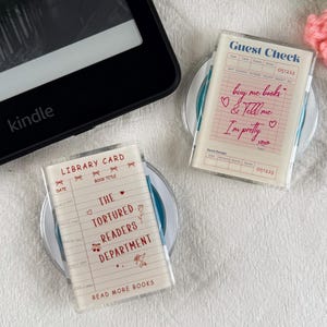 May include: Two clear plastic rectangular cases featuring printed designs. One displays a library card graphic with the text THE TORTURED READERS DEPARTMENT, while the other shows a guest check graphic with the text buy me books & Tell me I'm pretty.