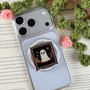 May include: A silver smartphone featuring a square phone grip with a dark, stamp-shaped illustration of a white ghost against a night sky with orange autumn leaves. The device is surrounded by a decorative green and pink crocheted vine.
