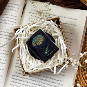 Puede incluir: Un pin rectangular con una ilustración estilo vidriera de un paisaje montañoso bajo un cielo nocturno con una gran luna. El pin tiene un borde negro y el texto VELARIS CITY OF STARLIGHT impreso en color dorado en la parte inferior.