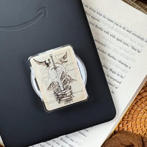 May include: A rectangular phone grip featuring a black and white ink-style illustration of a sword embedded in a stack of books, flanked by two roses, mountains, a moon, and two flying birds, attached to a black e-reader device.