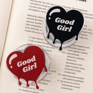 May include: Two heart-shaped phone grips with a glossy finish. One is black and the other is red, both featuring a white dripping effect at the bottom and the text Good Girl in white lettering. The designs are layered over a clear circular base.