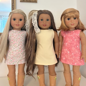 Puede incluir: Tres mu&ntilde;ecas de pie una al lado de la otra. La mu&ntilde;eca de la izquierda tiene el pelo largo y rubio y un vestido floral. La mu&ntilde;eca central tiene el pelo largo y casta&ntilde;o, un tocado de encaje blanco y un vestido amarillo. La de la derecha tiene un vestido rosa.