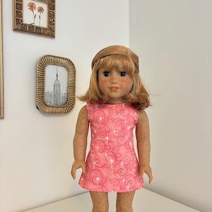 Puede incluir: Una mu&ntilde;eca con cabello casta&ntilde;o claro hasta los hombros y pecas, que viste un vestido rosa sin mangas con un patr&oacute;n de remolinos rosa oscuro y destellos blancos, combinado con botas de color canela. La mu&ntilde;eca tiene una expresi&oacute;n neutra.