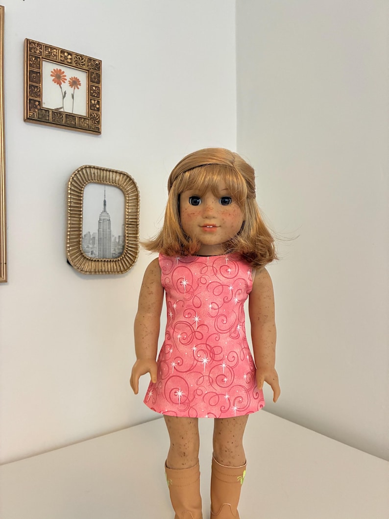 Puede incluir: Una mu&ntilde;eca con cabello casta&ntilde;o claro hasta los hombros y pecas, que viste un vestido rosa sin mangas con un patr&oacute;n de remolinos rosa oscuro y destellos blancos, combinado con botas de color canela. La mu&ntilde;eca tiene una expresi&oacute;n neutra.