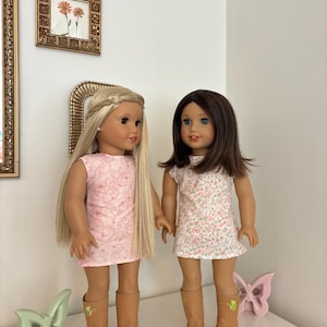 Puede incluir: Dos mu&ntilde;ecas de pie una al lado de la otra. La mu&ntilde;eca de la izquierda tiene el pelo largo y rubio con una diadema trenzada y lleva un vestido rosa con un patr&oacute;n de remolinos. La mu&ntilde;eca de la derecha tiene el pelo casta&ntilde;o y lleva un vestido floral.