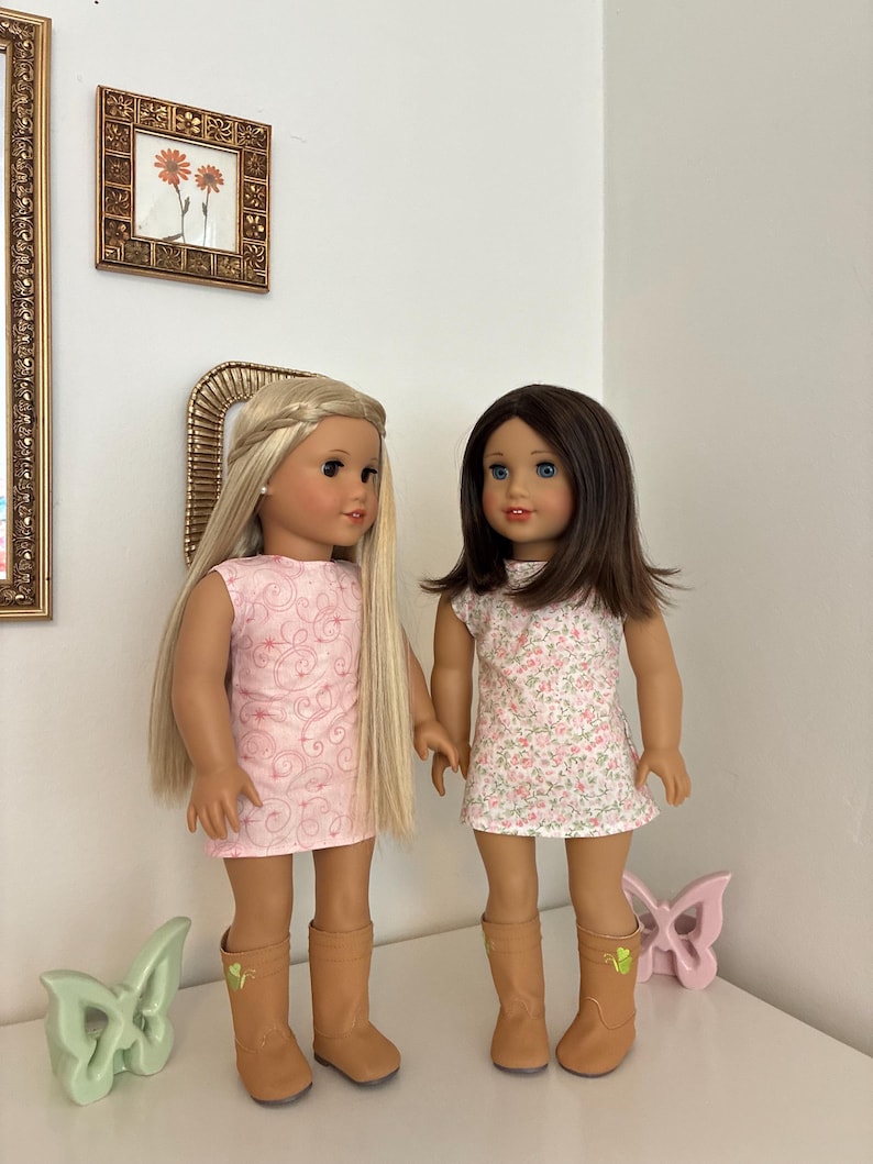 Puede incluir: Dos mu&ntilde;ecas de pie una al lado de la otra. La mu&ntilde;eca de la izquierda tiene el pelo largo y rubio con una diadema trenzada y lleva un vestido rosa con un patr&oacute;n de remolinos. La mu&ntilde;eca de la derecha tiene el pelo casta&ntilde;o y lleva un vestido floral.