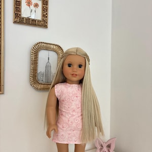 Puede incluir: Una mu&ntilde;eca con cabello rubio largo y liso peinado con una diadema trenzada, que viste un vestido rosa claro sin mangas con un delicado patr&oacute;n de remolinos rosa oscuro y botas de color tostado. La mu&ntilde;eca tiene ojos marrones y expresi&oacute;n neutra.