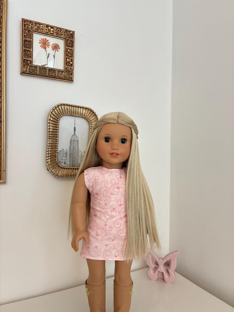 Puede incluir: Una mu&ntilde;eca con cabello rubio largo y liso peinado con una diadema trenzada, que viste un vestido rosa claro sin mangas con un delicado patr&oacute;n de remolinos rosa oscuro y botas de color tostado. La mu&ntilde;eca tiene ojos marrones y expresi&oacute;n neutra.
