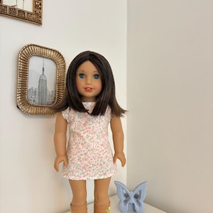 Puede incluir: Una mu&ntilde;eca con cabello casta&ntilde;o hasta los hombros y ojos azules, que viste un vestido blanco sin mangas con un peque&ntilde;o estampado floral rosa y botas de color canela. La mu&ntilde;eca tiene un tono de piel claro y una expresi&oacute;n neutra.