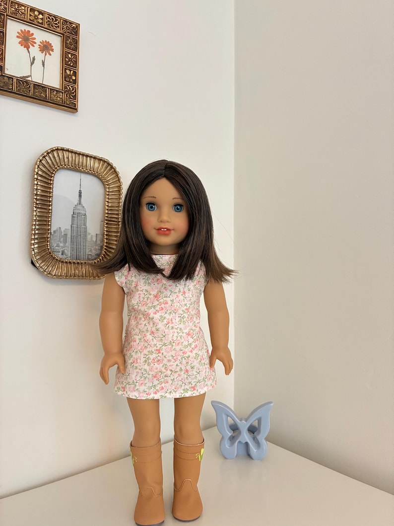 Puede incluir: Una mu&ntilde;eca con cabello casta&ntilde;o hasta los hombros y ojos azules, que viste un vestido blanco sin mangas con un peque&ntilde;o estampado floral rosa y botas de color canela. La mu&ntilde;eca tiene un tono de piel claro y una expresi&oacute;n neutra.