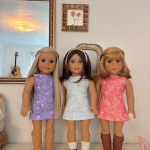 Puede incluir: Tres mu&ntilde;ecas de pie una al lado de la otra. La mu&ntilde;eca de la izquierda tiene el pelo largo y rubio, viste un vestido morado con estampado de espirales y botas de color canela. La mu&ntilde;eca central tiene el pelo casta&ntilde;o, una diadema blanca, un vestido azul claro con espirales, calcetines blancos y zapatos negros. La mu&ntilde;eca de la derecha tiene el pelo caoba, un vestido rosa con espirales y botas marrones.