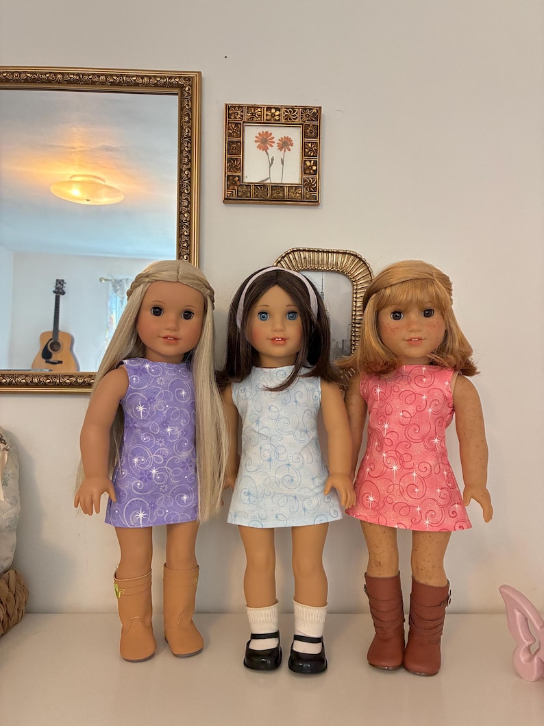 Puede incluir: Tres mu&ntilde;ecas de pie una al lado de la otra. La mu&ntilde;eca de la izquierda tiene el pelo largo y rubio, viste un vestido morado con estampado de espirales y botas de color canela. La mu&ntilde;eca central tiene el pelo casta&ntilde;o, una diadema blanca, un vestido azul claro con espirales, calcetines blancos y zapatos negros. La mu&ntilde;eca de la derecha tiene el pelo caoba, un vestido rosa con espirales y botas marrones.
