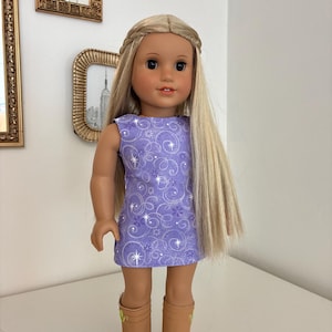 Puede incluir: Una mu&ntilde;eca con cabello rubio largo peinado con una diadema trenzada, que viste un vestido morado sin mangas con un patr&oacute;n de estrellas y flores blancas en espiral, combinado con botas color canela hasta la pantorrilla con detalles verdes.