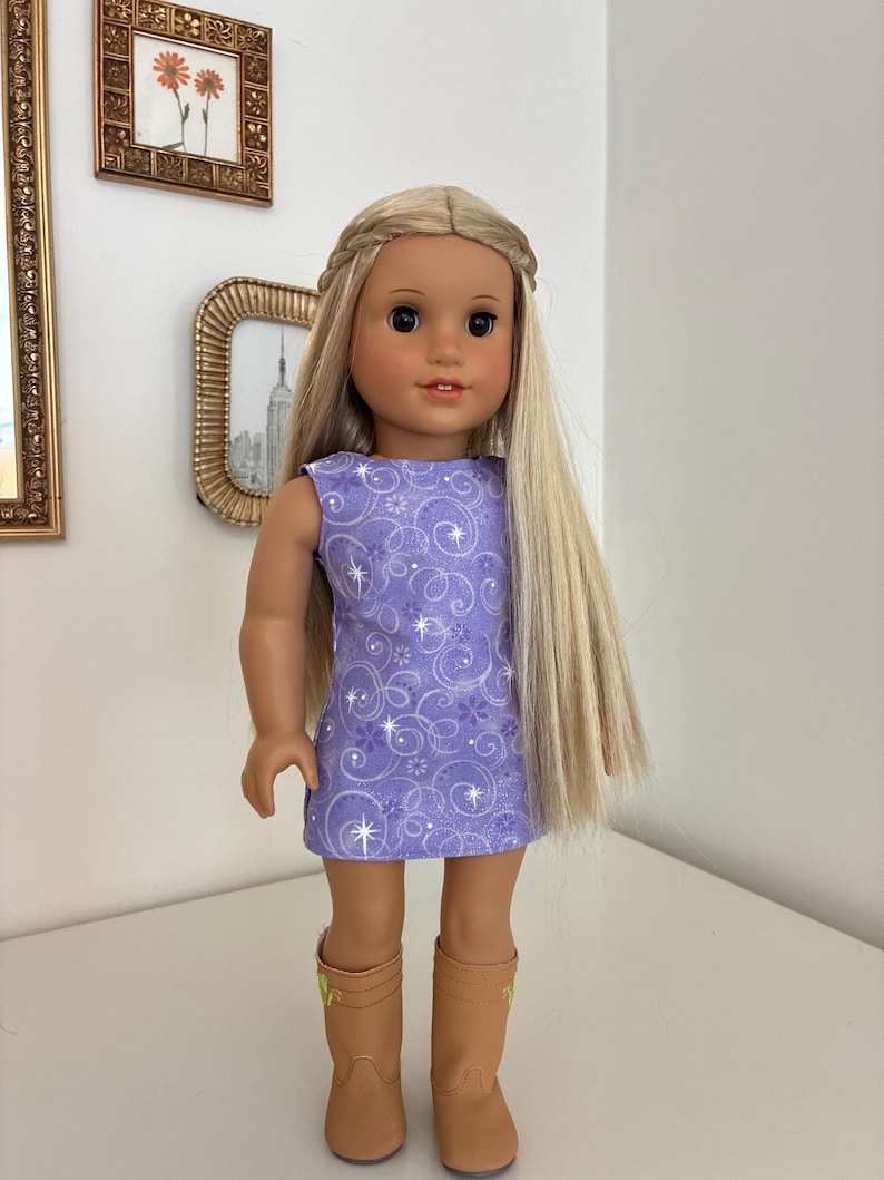 Puede incluir: Una mu&ntilde;eca con cabello rubio largo peinado con una diadema trenzada, que viste un vestido morado sin mangas con un patr&oacute;n de estrellas y flores blancas en espiral, combinado con botas color canela hasta la pantorrilla con detalles verdes.