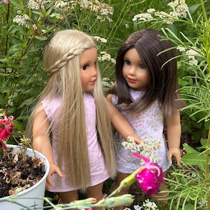 Puede incluir: Dos mu&ntilde;ecas de pie una al lado de la otra. La mu&ntilde;eca de la izquierda tiene el pelo largo y rubio con una corona trenzada y viste un vestido rosa claro. La mu&ntilde;eca de la derecha tiene el pelo casta&ntilde;o y viste un vestido blanco con flores.