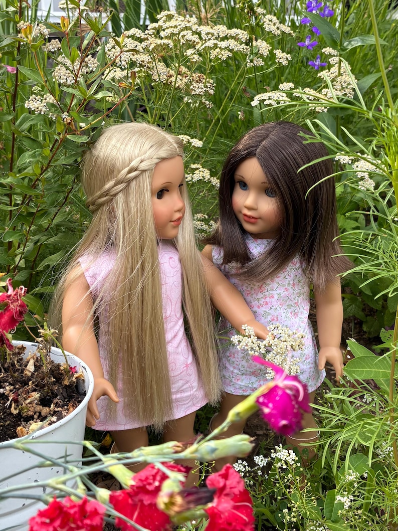Puede incluir: Dos mu&ntilde;ecas de pie una al lado de la otra. La mu&ntilde;eca de la izquierda tiene el pelo largo y rubio con una corona trenzada y viste un vestido rosa claro. La mu&ntilde;eca de la derecha tiene el pelo casta&ntilde;o y viste un vestido blanco con flores.