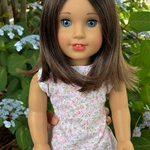 Puede incluir: Una mu&ntilde;eca con cabello casta&ntilde;o hasta los hombros, ojos azules y mejillas sonrosadas. La mu&ntilde;eca viste un vestido blanco sin mangas con un peque&ntilde;o estampado floral rosa y verde. La mu&ntilde;eca tiene una expresi&oacute;n facial neutra con dientes visibles.