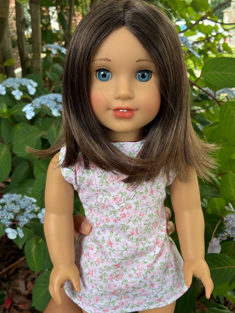 Puede incluir: Una mu&ntilde;eca con cabello casta&ntilde;o hasta los hombros, ojos azules y mejillas sonrosadas. La mu&ntilde;eca viste un vestido blanco sin mangas con un peque&ntilde;o estampado floral rosa y verde. La mu&ntilde;eca tiene una expresi&oacute;n facial neutra con dientes visibles.