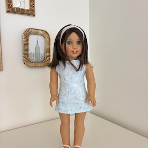 Puede incluir: Una mu&ntilde;eca con cabello casta&ntilde;o y ojos azules que viste un vestido sin mangas azul claro con un patr&oacute;n de remolinos blancos, una diadema rosa claro y zapatos rosa claro a juego. La mu&ntilde;eca tiene tez clara y est&aacute; de pie.