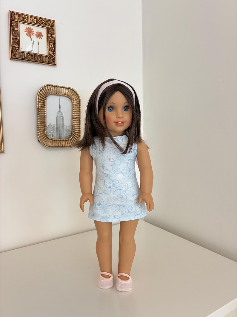 Puede incluir: Una mu&ntilde;eca con cabello casta&ntilde;o y ojos azules que viste un vestido sin mangas azul claro con un patr&oacute;n de remolinos blancos, una diadema rosa claro y zapatos rosa claro a juego. La mu&ntilde;eca tiene tez clara y est&aacute; de pie.