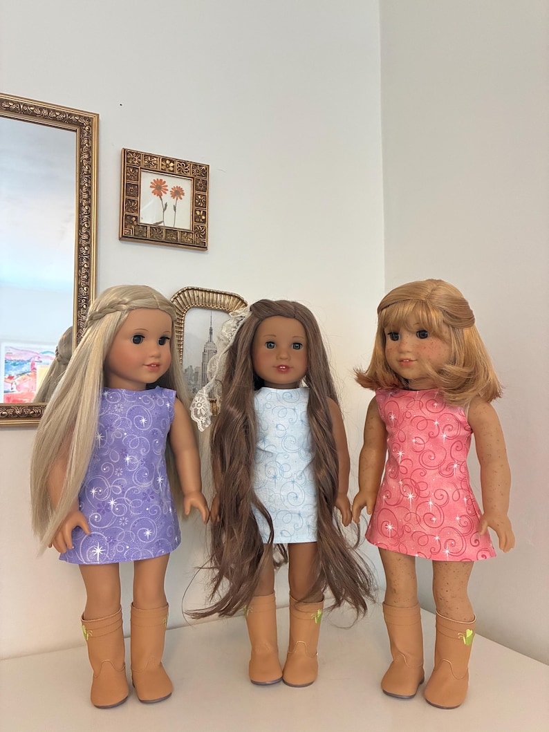 Puede incluir: Tres mu&ntilde;ecas de pie una al lado de la otra, cada una con un vestido sin mangas con un patr&oacute;n de remolinos y detalles de estrellas. La mu&ntilde;eca de la izquierda tiene el pelo largo y rubio, la del centro tiene el pelo largo y casta&ntilde;o, y la de la derecha tiene el pelo casta&ntilde;o rojizo hasta los hombros. Las tres llevan botas de color tostado.