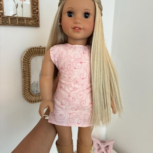 Puede incluir: Una mu&ntilde;eca con cabello rubio largo y liso peinado con una diadema trenzada, que viste un vestido rosa claro sin mangas con un patr&oacute;n de finos remolinos rojos y peque&ntilde;as estrellas, combinado con botas color canela hasta la pantorrilla.