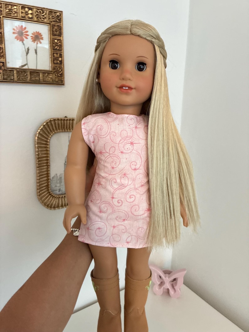 Puede incluir: Una mu&ntilde;eca con cabello rubio largo y liso peinado con una diadema trenzada, que viste un vestido rosa claro sin mangas con un patr&oacute;n de finos remolinos rojos y peque&ntilde;as estrellas, combinado con botas color canela hasta la pantorrilla.