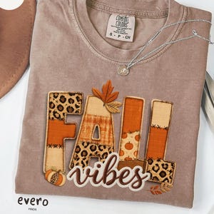 Puede incluir: Una camiseta de manga corta color espresso con el texto FALL VIBES. La palabra FALL está compuesta por letras de bloque con varios patrones que incluyen estampado de leopardo, cuadros y motivos de calabazas. Una hoja naranja sobre la A.