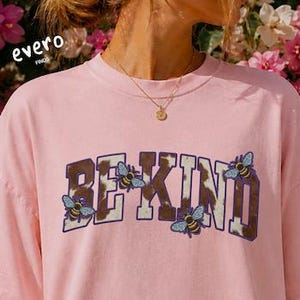 May include: A light pink short-sleeve t-shirt featuring the text BE KIND in a brown and white cow print pattern, accented with small illustrated bees. The shirt includes the text evero and blossum printed in small white circles.