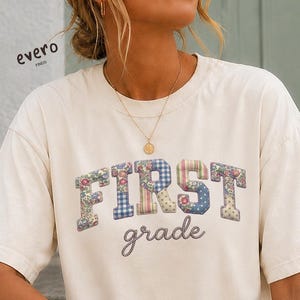 Puede incluir: Una camiseta de manga corta color marfil con la palabra FIRST en letras mayúsculas grandes con estampados florales y de cuadros vichy multicolores, y la palabra grade escrita debajo en cursiva gris oscuro. Tiene cuello redondo.