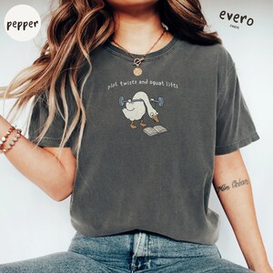 May include: A charcoal gray short-sleeve t-shirt featuring a graphic of a white goose lifting a barbell while reading an open book. The text above the goose reads, plot twists and squat lifts. The shirt has a crew neckline and a relaxed fit.