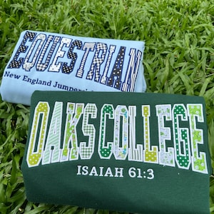 May include: A light blue sweatshirt with the text EQUESTRIAN New England Jumpers in dark blue patterned letters and a dark green sweatshirt with the text OAKS COLLEGE ISAIAH 61:3 in white-bordered letters featuring various green floral patterns.
