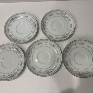 May include: A set of five white porcelain saucers featuring a delicate gray floral pattern along the rim. Each saucer has a central circular indentation for a cup and a thin silver-toned metallic trim around the outer edge of the plate.