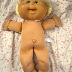 May include: A doll with a soft peach-colored fabric body, a vinyl head with blonde yarn hair, and green eyes. The doll features a wide, smiling mouth with two visible teeth, a small belly button, and a yellow bonnet with white lace trim.