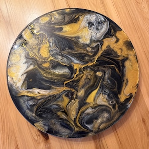 May include: A circular wall art piece featuring a glossy, poured resin finish with a marbled design. The surface displays a swirling pattern of metallic gold, deep black, and shimmering silver pigments that blend together in an abstract composition.