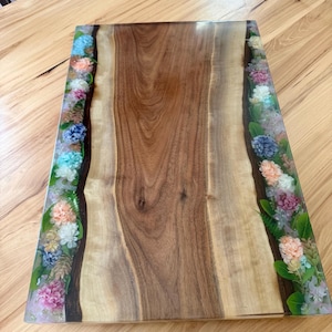 May include: A rectangular wooden serving board featuring a central natural wood grain panel flanked by two clear resin borders. Each resin section is embedded with a variety of colorful dried flowers and green leaves in shades of blue, pink, and white.