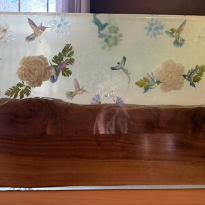 May include: A rectangular decorative block featuring a natural wood base with a live edge and a clear resin top. The resin section is embedded with pressed flowers, green foliage, and multiple colorful hummingbird illustrations.