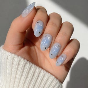 Peut inclure: Ongles de forme ovale avec une base bleu clair, ornés d'étoiles blanches et de petits strass transparents. L'ongle du pouce présente un motif de soleil blanc, tandis que les autres ongles sont décorés d'étoiles et de pierres scintillantes.