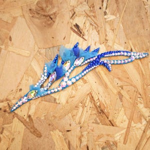 May include: A decorative hair accessory featuring a bright blue base adorned with numerous sparkling clear and iridescent rhinestones, small dark blue beads, and layered blue tulle fabric accents arranged in a curved, elongated branch-like shape.