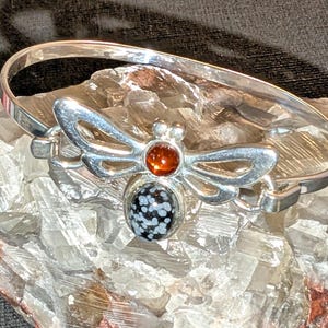 Può includere: Un bracciale rigido in metallo color argento con un ornamento centrale a forma di insetto. L'insetto presenta una pietra liscia, rotonda e bruno-rossastra per la testa e una pietra nera ovale più grande con inclusioni bianche per il corpo.