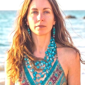 May include: A multi-strand necklace featuring numerous round, bright turquoise beads strung together in a layered design, accompanied by matching turquoise bead drop earrings with silver-toned hardware, worn with a patterned teal and maroon top.