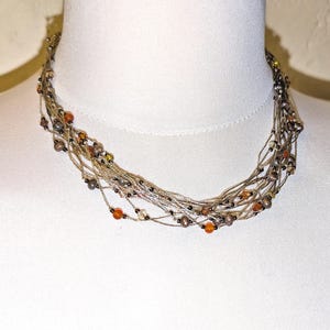 May include: A multi-strand necklace with a neutral color palette. The necklace features numerous strands of thin, silver-colored cords, adorned with small, colorful beads in shades of orange, brown, and black. The necklace is displayed on a white mannequin.
