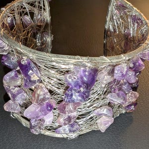May include: A silver wire cuff bracelet, embellished with polished amethyst stones. The bracelet has a woven wire design, with the purple gemstones clustered around the edges. The amethyst stones provide a striking contrast to the silver.