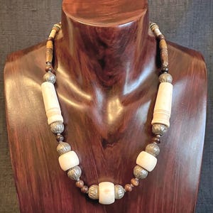 May include: A necklace featuring a sequence of cylindrical and spherical beads. The design alternates between smooth cream-colored cylindrical beads, smaller dark brown wooden beads, and textured metallic silver-toned spherical spacers.