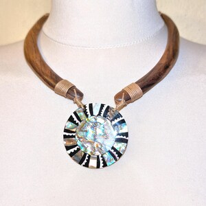 May include: A necklace featuring a curved, polished brown wooden collar connected by tan cord wrappings to a circular pendant. The pendant displays a central iridescent abalone shell inlay surrounded by alternating black and white scalloped segments.