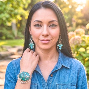 May include: A woman wearing turquoise earrings and a matching bracelet. The earrings have a silver-coloured metal design with turquoise beads. The bracelet features a large turquoise stone wrapped in silver-coloured wire. She is wearing a denim shirt.